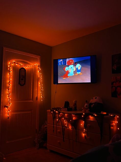 Orange lights decorating a room giving it a Halloween theme with jack the skeleton squishmallow and snoopy dressed as a pumpkin stuffed animal. Trick or treat mickey mouse special playing on tv. Halloween Lights Bedroom, Halloween Theme Room, Halloween Room Ideas, Halloween Themed Room, Halloween Lights Indoor, Trick Or Treating Aesthetic, Cozy Halloween Bedroom, Fall Room Decorations, 200 Aesthetic