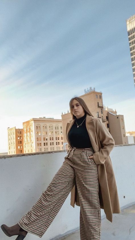 Girl standing on a city parking structure wearing beige plaid wide leg pants, black shirt, long tan overcoat and brown heeled boots. Tan Pants Outfit Winter, Tan Trousers Outfit Women, Plaid Pants Outfit Winter, Tan Trousers Outfit, Plaid Trousers Outfit, City Winter Outfit, Tan Pants Outfit, Tan Overcoat, Plaid Wide Leg Pants