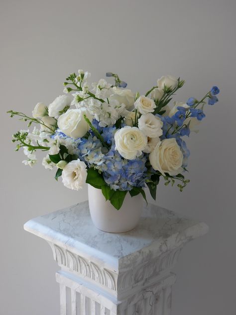 An Elegant White and Blue Floral Arrangement Introducing our Sky Blue arrangement—a stunning floral display featuring an elegant selection of white and blue flowers and lush foliage, artfully arranged in a low cylinder glass vase. This exquisite bouquet includes ranunculus, hydrangeas, delphinium, lisianthus, roses, and more, creating a harmonious blend of colors reminiscent of clear skies and tranquil waters. The Sky Blue arrangement embodies serenity and sophistication, making it the perfect c Blue White Green Floral Arrangements, Sky Blue And White Bouquet, Blue White Flower Arrangements, Blue Hydrangea Arrangements, Hydrangea Vase Arrangement, Blue Flower Decorations, Blue And White Flower Arrangements, White And Blue Floral Arrangements, Blue And White Floral Arrangements