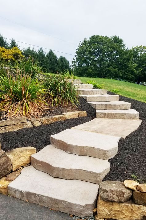 We designed and installed these stone steps that connected a driveway to an upper porch entrance area. Built into the existing slope of a hill, the landing areas between the steps incorporate smaller pavers with a stylish laying pattern. Steps and walkways are landscape elements that offer a great chance to express your unique style and enhance nature's beauty. Landscaping Stone Steps, Sloped Front Yard Steps, Landscape Ideas For Sloping Backyard, Landscaping Steps On A Hill, Landscape Steps On A Hill, Natural Steps On Hill, Patio Built Into Hill, Outdoor Landscape Steps, Garden Steps Ideas On A Slope