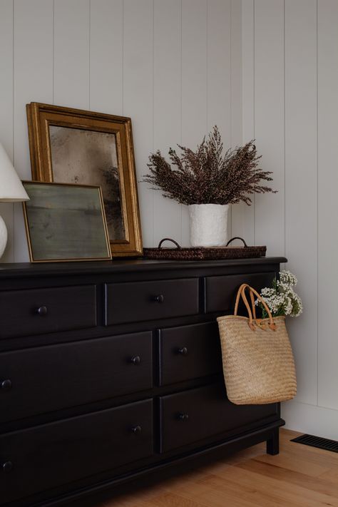 Cabin and california casual inspired bedroom. Black dresser with black knobs paired with a gold vintage mirror and dried florals. Vertical planks in a bedroom with a wood planked ceiling. Neutral bedroom decor. Putty white wall inspiration. Straw market bag styling. Bedroom styling by Nadine Stay | #bedroomdecor #bedroomdresser #blackdresser #californiacasual #bedroomstyling #bedroomdecor #verticalplanks #shiplap #verticalpanels #plankedwalls Bedroom Black Dresser, Wood Planked Ceiling, California Casual Bedroom, Gold Vintage Mirror, Planked Ceiling, Bedside Table Styling, Styling Bedroom, Casual Bedroom, Vintage Gold Frame