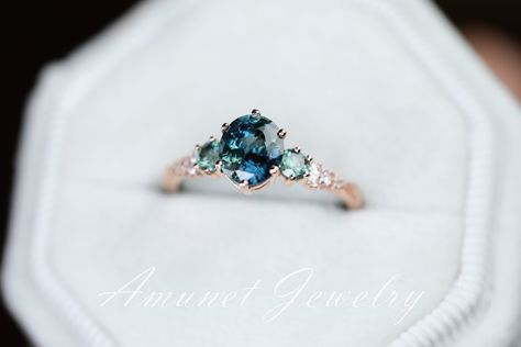 This engagement ring has a stunning peacock sapphire. Its beauty is enhanced by the sparkle of two 3mm round light green Montana sapphires, sparkling white diamonds cluster and leaves on the gold band on either side.The ring can be ordered solo or with lovely matching band in 14K rose, yellow or white gold.The detailed Peacock Sapphire Ring, Peacock Sapphire Engagement Ring, Diamond And Sapphire Engagement Ring, Sapphire Ring Vintage, Peacock Sapphire, Blue Green Sapphire Ring, Sapphire Cluster Ring, Oval Sapphire Engagement Ring, Montana Sapphires