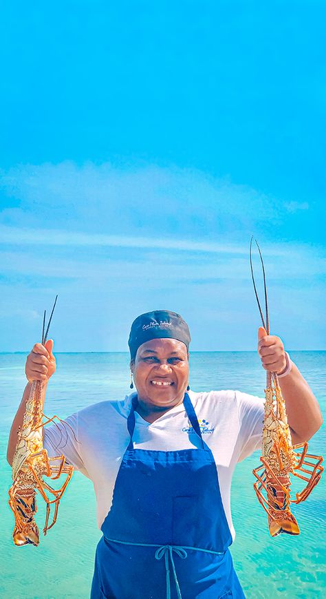 Ms. Jude - Chef from Coco Plum Island Resort holding a lobster in each hand. With the beautiful Caribbean Sea in the background and a blue sky. Lobster Bib, Lobster Fest, Grilled Lobster, Plum Island, Belize Travel, Seasonal Celebration, Live Entertainment, Private Island, Island Resort