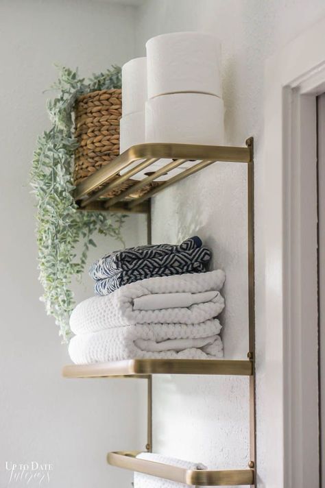 Glam Bathroom Decor with a gold towel bar and shelves housing towels, toilet paper, and a plant on white walls.  Find other ideas for a small bathroom makeover on a budget.   #smallbathroomideas #smallbathroomideasonabudget #bathroomdecorideas #bathroomideasonabudgetdiy #bohobathroom Bathroom Shelves Over Toilet For Towels, Gold Bathroom Shelves Over Toilet, Metal Shelves Bathroom, Vanity Shelf Ideas Bathroom, Small Bathroom Simple Ideas, Gold Shelves Bathroom, Towels Over Toilet, Simple Chic Bathroom, Bathroom Decor Above Towel Bar