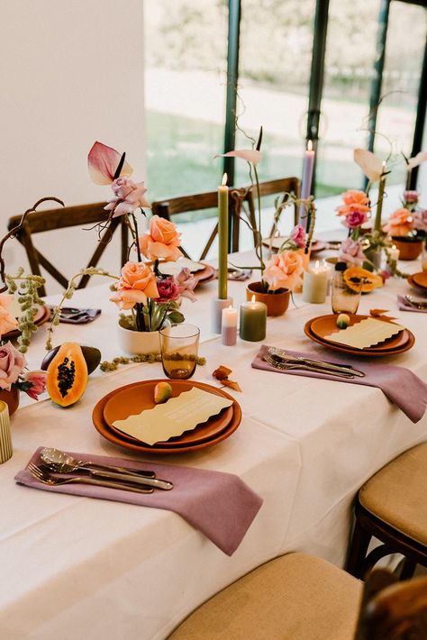 Terracotta wedding plates with lavender wedding napkins, fruit decor, and green and purple candles Colored Candles Wedding Table, Terracotta Lavender Wedding, Colored Candlesticks Wedding, Terracotta And Lavender Wedding, Purple And Terracotta Wedding, Green And Purple Wedding Decor, Colorful Tablescapes Wedding, Green And Orange Centerpieces, Lavender Orange Green Wedding