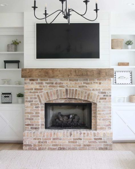 This white shiplap wall sports a coordinating white framed TV above a rough-hewn unfinished wood mantel and weathered brick fireplace. White built-in shelves and cabinets on either side display white, black, and wood decorations, while a modern black metal light fixture hangs from the ceiling in the center of the room. Fun Fireplace, Fireplace Wall Ideas With Tv, Brick Fireplace White, White Stone Fireplaces, Stone Fireplace Wall, White Brick Fireplace, Faux Fireplace Mantels, Chicago Brick, Tv Over Fireplace