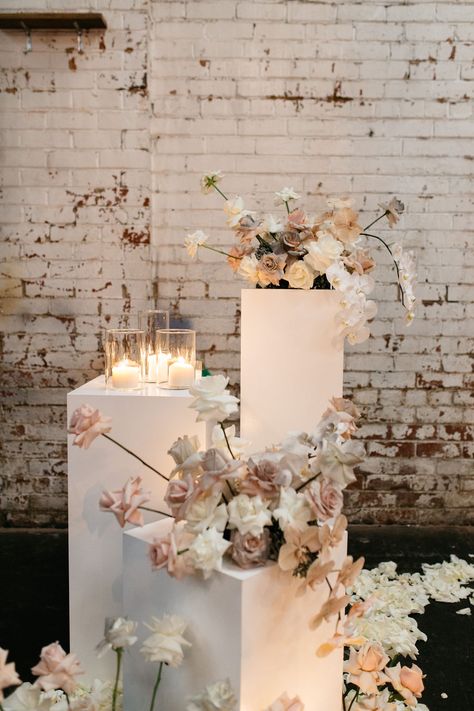 White wedding plinths topped with soft pink roses and cascading phalenopsis orchids are highlighted by clusters of romantic pillar candles in glass pillar candle holders. This makes a striking contrast to the industrial warehouse venue and rustic brick wall backdrop. Styling by: Ruffles and Bells candles by Southern Lights Candle Co. Wedding Pillars, Romantic Florals, Ceremony Inspiration, Ceremony Flowers, Ceremony Backdrop, Wedding Aisle, Wedding Ceremony Decorations, Wedding Mood, White Vases