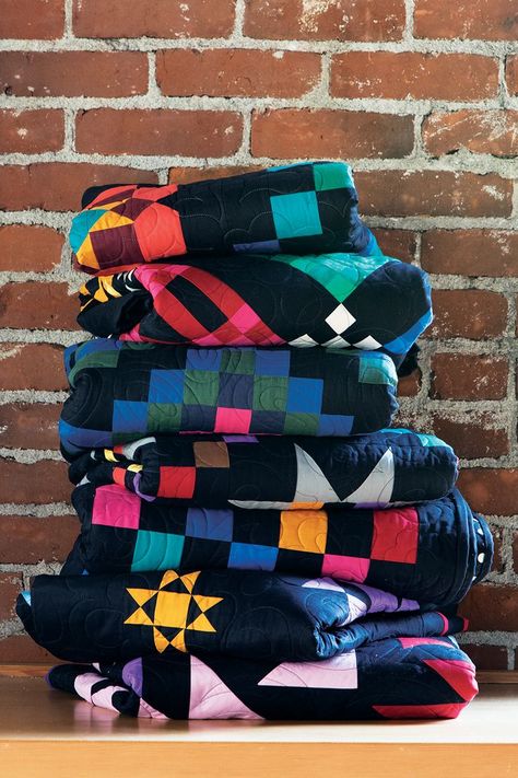 A stack of folded quilts are in front of a red brick wall. The quilts all feature black backgrounds and colorful patterns. Amish Quilt Patterns, History Of Quilting, Amish Quilt, Geometric Patchwork, Connecting Threads, Lemon Chiffon, Amish Quilts, Amish Recipes, Sampler Quilt