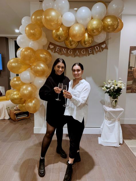 Two Indian women standing in front of decoration display made up or gold and white balloons and a banner that says engaged on it, cheersing with champagne glasses Simple Engagement Party Ideas At Home, Casual Engagement Party Decorations, Small Engagement Party Ideas Simple, Gold Engagement Party Decor, White And Gold Engagement Party, Engagement Party Decorations Indoor, At Home Engagement Party, Elegant Engagement Party Ideas, Classy Engagement Party