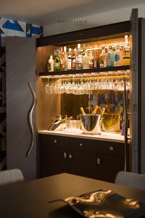 The rich flamed oak of this bar has been angled alternately between panels, giving a chevroned detail that adds subtle character to the piece, whilst overall remaining understated in the dining room. The eye-catching fluid cast aluminum handles and bespoke gold on bronzed mirror backsplash complement the ebonised tones in the wood for a classic, industrial feel. Mirror Wall Bar Design, Bar With Mirror Backsplash And Lighting, Mirror Bar Shelves, Home Bars With Mirrors, Bar Mirror Backsplash, Mirror Wall Bar, Bar With Mirror Backsplash, Bar With Mirror, Mirrored Bar