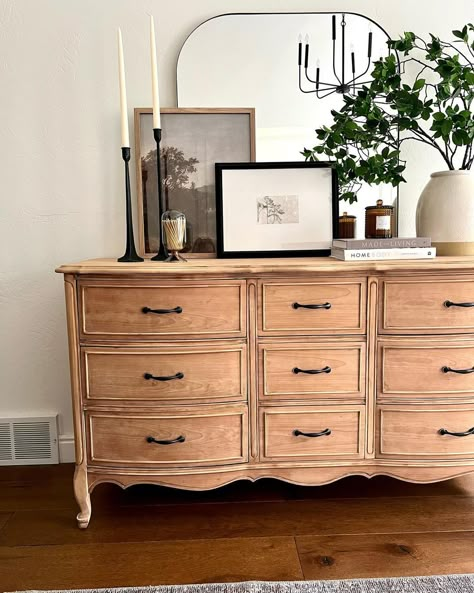 A vintage wood dresser with vintage black handles is pushed against a white bedroom wall. A large plant in a white pot, a stack of two books, a couple of pictures, and two black candlesticks with white candles are placed in front of a large mirror on the dresser. A grey rug is laid on the warm wood floor. Wardrobe Decoration, Dresser Styling, Room Wall Decor Bedroom, Dresser Decor Bedroom, Black Candlesticks, Warm Wood Flooring, Bedroom Wall Decor Ideas, White Wall Bedroom, Black Dresser