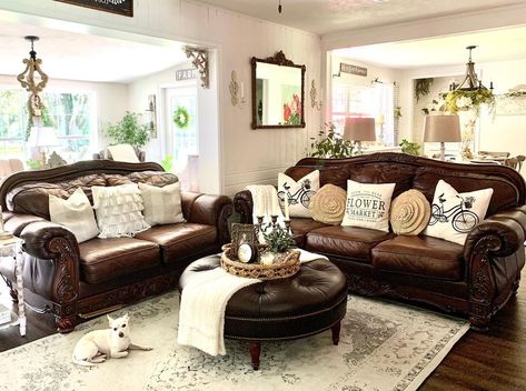 This farmhouse living room boasts a brown leather couch with antique dark wood frame seated next to a matching loveseat. A round tufted brown leather ottoman is placed in front of the couch on a beige and gray vintage rug. Styling A Dark Leather Couch, Farmhouse Living Room Brown Leather Sofa, Brown Loveseat Living Rooms, Decorating With Dark Brown Leather Couch, Living Room With Dark Brown Leather Furniture, Dark Brown Leather Sofa Living Room Decorating Ideas, Farmhouse Living Room With Leather Couch, Rugs To Match Brown Leather Couch, Living Rooms With Brown Leather Couches