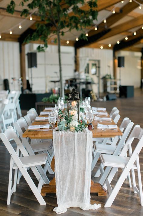 Wedding reception long wooden tables with white draped table runners, lanterns, and white red and greenery centerpieces with twinkle lights at wedding. #weddingreception #rusticweddingdecor #weddingdecor #weddingtablescape White Table Runners Wedding, Wood Tables With White Chairs Wedding, Lantern Centerpieces Long Table, Wooden Table Decor Wedding, Wooden Tables Wedding, Wooden Table Wedding Decor, White Chairs Wedding, Lobola Decor, Wedding Ideas 2024