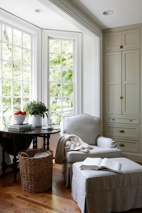 In a kitchen bay window nook, a light gray skirted chair and ottoman sit beside a dark stained wooden table and a wicker basket. #windows #homedecor #interiordesign #decoration Taupe Paint Colors, Kitchen Bay Window, Taupe Paint, White Cabinetry, Amber Interiors, Plywood Furniture, Reading Nooks, Style At Home, Interior Design Firms