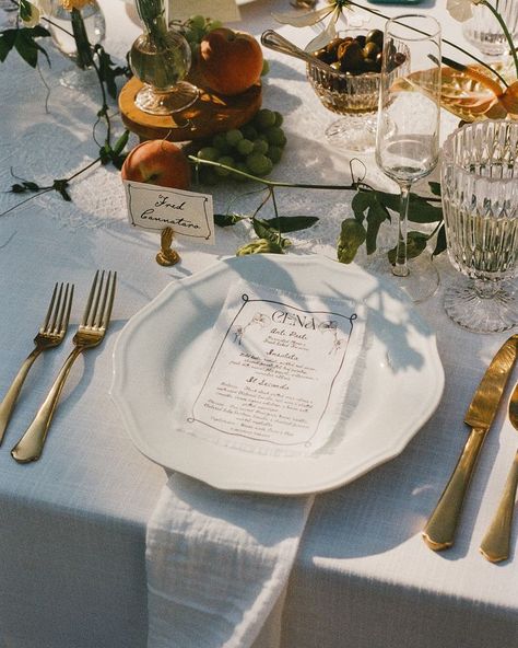 Elegant wedding place setting with white plates, gold cutlery, crystal glasses, and Italian menu card decorated with fresh fruit and greenery Wedding Tablescape Simple, Italian Dinner Table Setting, Italian Wedding Table Setting, Mediterranean Wedding Aesthetic, Reception Dinner Table Set Up, Table Settings Italian, Fruit Tablescape Wedding, Wedding Plate Settings, Medditeranean Wedding