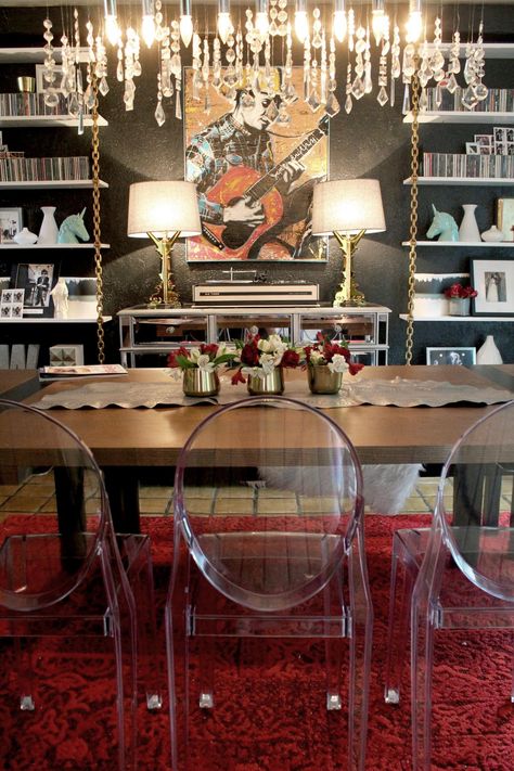 This eclectic dining room features white bookshelves with chains, a mirrored buffet table and two gold table lamps positioned in front of a black wall. A long crystal chandelier is positioned above a rectangular wooden dining table, while Louis XIV ghost chairs and a deep red area rug finish the look of the neutral space. Rock And Roll Room, Dream Dining Room, Eclectic Dining Room, Dining Room Accents, Eclectic Dining, Dining Room Lamp, Storage Inspiration, Mid Century Modern Living Room, Gold Table Lamp