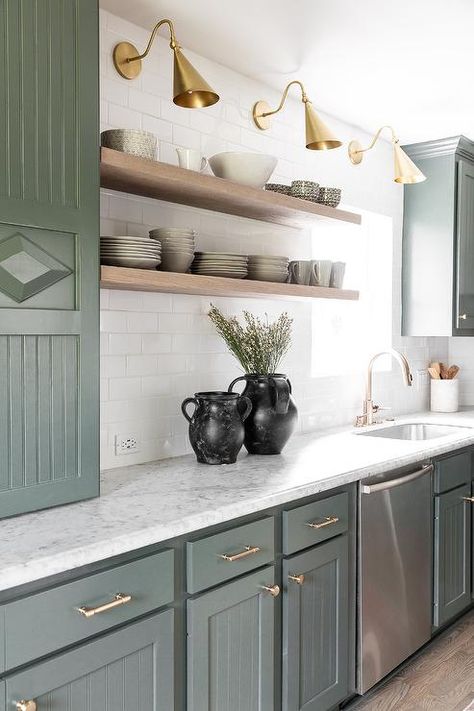 Green and white kitchen features green kitchen beadboard cabinetry with vintage brass pulls and honed carrera gray marble countertops and stacked floating brown shelves mounted on white subway wall tiles illuminated by brass sconces. Illusive Green, Gray Marble Countertops, Green Beadboard, Beadboard Cabinets, Kitchen Beadboard, White Cottage Kitchens, Brown Shelves, Corner Stove, Green Kitchen Island