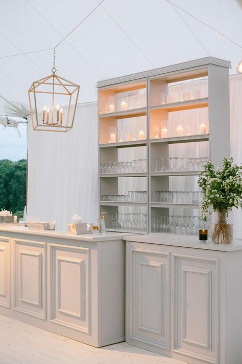 Create a sophisticated ambiance with this elegant white wedding bar setup. Featuring classic cabinetry and a beautifully arranged shelf with glasses and candles, this bar is perfect for serving drinks in style. The clean, minimalist design is complemented by a geometric lantern and lush greenery, making it an ideal addition to any upscale wedding reception. #WeddingBar #ElegantWedding #WeddingReception #WeddingDecor #SophisticatedStyle Wedding Bar Setup, Easton Events, Wedding Bars, Bars Design, Mesa Bar, Elegant White Wedding, Food Counter, Kt Merry, Next Luxury