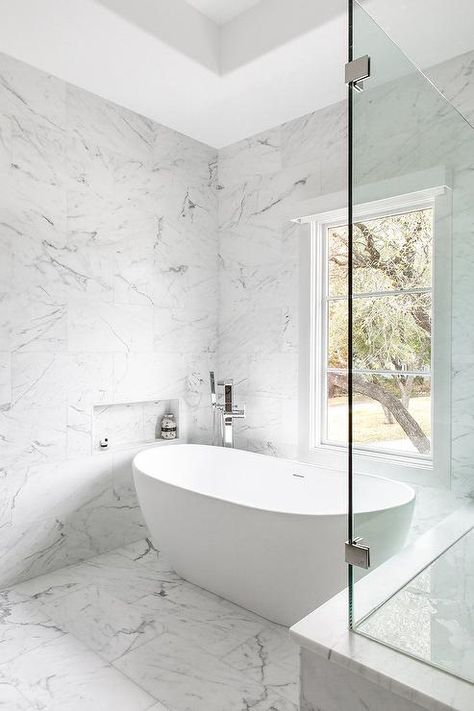 A marble tiled niche, surrounded by marble wall tiles, is positioned above an oval freestanding bathtub paired with a polished nickel modern floor mount tub filler and placed on marble floor tiles beneath a window. Tiled Niche, Bathroom Tub Remodel, Tile Shower Niche, Tub Remodel, Bathroom Freestanding, Marble Tile Bathroom, Marble Wall Tiles, Sophisticated Bathroom, Marble Tile Floor