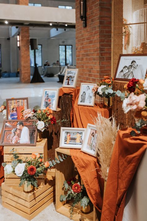 A mini photo display at the wedding reception clad in a burnt orange theme, decorated with wooden elevations and floral arrangements. Rusty Orange Wedding Decor, Burnt Orange Spring Wedding, Red And Burnt Orange Wedding, Burnt Orange Table Decor, Burnt Orange Wedding Centerpieces, Green And Orange Wedding Theme, Black And Burnt Orange Wedding, Burnt Orange Wedding Decor, Rusty Orange Wedding