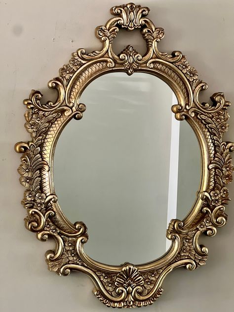 Iconic in its design and reminiscent of Old-Hollywood glamour, this ornately carved, oval, 1960s gold gilt mirror glams up your wall lending an inherent sense of luxury to any space.  Good vintage condition with gilt having acquired a warm patina. No cracks of any kind. Gorgeous Gold Gilt Heavy Plastic Composite Frame. Some visible shading and wear of gold gilt paint on frame and outer sides, which only adds glamour to the warm patina. One clamp for hanging the mirror is broken but the mirror ca Vintage Mirrors Small, Circle Vintage Mirror, Ornate Wall Mirror, French Gold Mirror, Oval Vintage Mirror, Gold Apartment Aesthetic, Vintage Gold Decor, Gold Aesthetic Home Decor, Mirror In Office