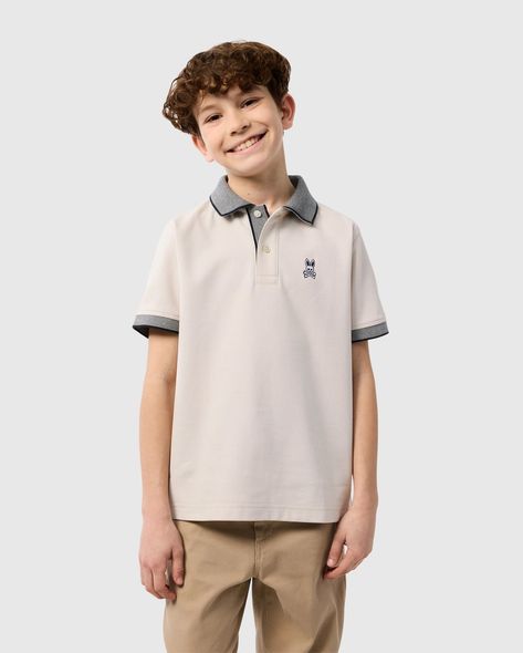 A smiling young boy with curly brown hair is wearing a beige KIDS SOUTHPORT PIQUE POLO SHIRT - B0K263B200 by Psycho Bunny made of Pima cotton piqué with grey collar and sleeve trim, and khaki pants. He stands against a plain light grey background. Polo Shirt Outfit Men, Polo Shirt Outfits, Polo Fashion, Shirt Outfit Men, Boys Tops, Polo Tees, Swimwear Shorts, Pique Polo Shirt, Boys Top