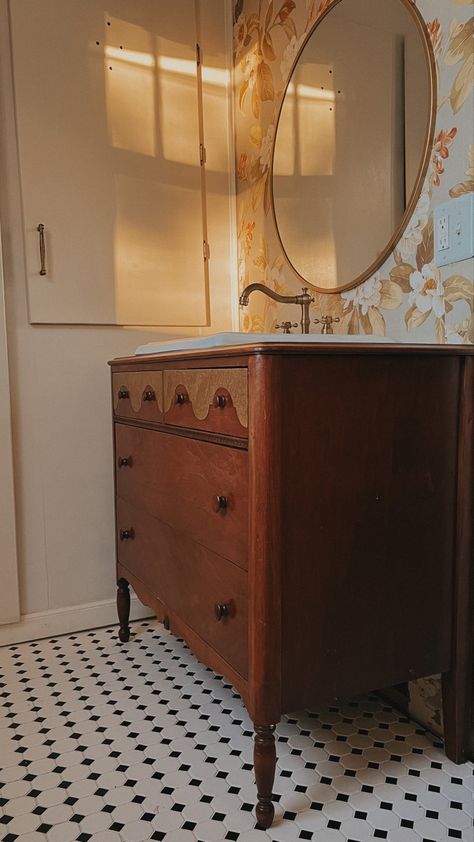 Wooden dresser, turned into a bathroom sink, vanity with floral wallpaper in the background, brass hardware, and a black and white checkered floor Vintage Wooden Vanity Bathroom, Making A Dresser Into A Bathroom Vanity, Old Dresser To Bathroom Vanity, Dresser As Vanity Bathroom, Dresser To Sink Vanity, Vintage Dresser Bathroom Vanity, Old Dresser Bathroom Vanity, Dresser As Vanity, Antique Dresser Bathroom Vanity