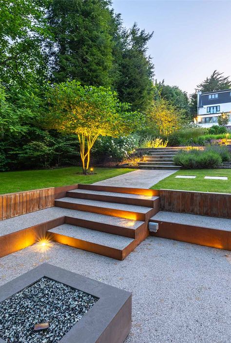 This contemporary garden is a combination of design and functionality. Tiered to complement the natural slope of the landscape, it's the perfect setting for both entertaining and family living.  This tier has been designed as an entertainment space around the unique fire pit   #LandscapeDesign  #GardenInspiration #OutdoorEntertaining #SurreyGardens #GardenMaintenance #slopedGarden idea #TieredGarden #DesignIdea  #GardenInspo #GardenIdeas #GardenDesign #Countrygarden #FirePit #Gardenlighting Garden Design Tiered, Garden On Different Levels, Garden Multi Level, 3 Level Garden Ideas, 3 Tier Garden Ideas, Two Tier Patio, Different Level Garden Ideas, Two Tier Garden Ideas, Tiered Landscaping Sloped Backyard