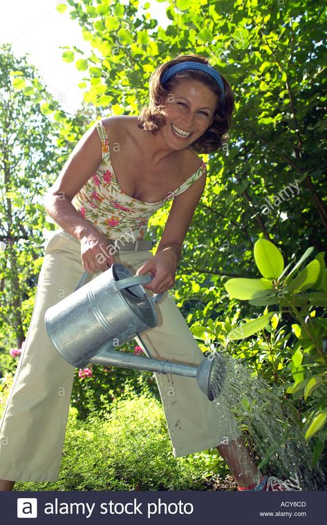 smiling woman in garden watering some plants with a watering can Stock Photo Holding Watering Can Reference, Person Watering Plants Reference, Watering Plants Pose Reference, Watering Plants Reference, Person Watering Plants, Woman Watering Plants, Fantasy Cafe, Woman In Garden, Irl References