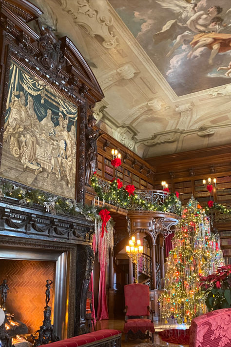 Inside the Biltmore Estate, the library radiates holiday charm. A tall Christmas tree with colorful lights and tinsel stands near red velvet chairs. The grand fireplace, framed by intricate wood carvings and topped with a tapestry, glows warmly. A spiral staircase that is led to garlands with red bows that drapes over the mantle and balcony, while the ceiling features a stunning mural of angels and clouds, adding a majestic touch to the festive, book-filled room. Mansion Library Aesthetic, Biltmore Estate Library, Biltmore House Christmas, Estate Library, Mansion Library, Biltmore Estate Interior, Biltmore Estate Christmas, Staircase Fireplace, Biltmore Christmas