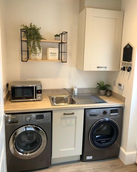 This multifunctional utility room is far from boring thanks to a steel gray washer and dryer fitted under a light wood counter. A stainless steel sink is built into the counter next to a stainless microwave. Create plenty of storage space with off-white cabinets and light wood and black metal shelves. Laundry Room With Microwave, Stainless Steel Laundry Room, Small Utility Room With Window, Washer Dryer Under Sink, Microwave In Laundry Room, Sink Between Washer And Dryer, White Utility Room, Utility Room With Sink, Counter Over Washer And Dryer