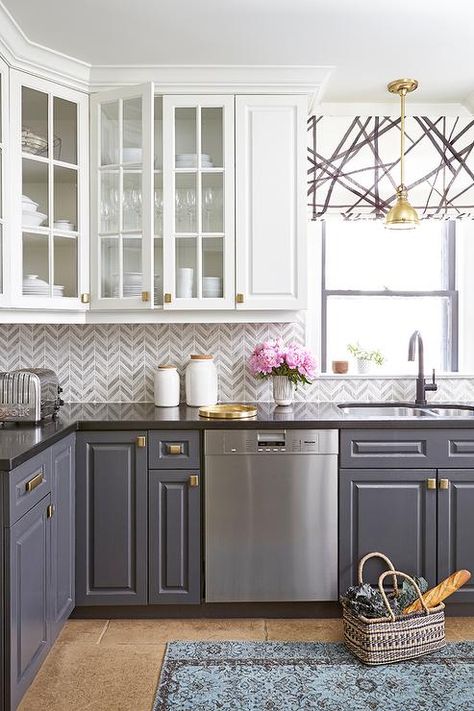 Stunning kitchen features white upper cabinets and gray lower cabinets adorned with brass hardware paired with black quartz countertops and white and gray marble chevron backsplash tiles. Two Tone Kitchen Cabinets, Серая Кухня, Decorative Shelving, Kabinet Dapur, Herringbone Backsplash, Two Tone Kitchen, Dream Kitchens, Kitchen Cabinets Makeover, Grey Kitchen Cabinets