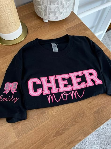 ** Please read before buying **  Personalized Embroidered Cheer Mom Shirt with glitter HTV applique. Fit: Unisex, Fits loose/relaxed. Size up for a more oversized fit.  Sizing: S,M,L,XL,2XL Material: 100% Cotton Embroidery Details: "Cheer Mom" centered across the chest Bow and Child name on right shoulder sleeve PROCESSING/TURNAROUND TIME: -Processing/Turn around times may vary. Please check the estimated ship/delivery times. -Processing/Turn around time DOES NOT include shipping time. ** Please note the picture is a sample only & it will be made once the order is placed. Colors will vary slightly due to lighting & device settings ** CARE INSTRUCTIONS: Machine wash cold inside out. Lay flat to dry or tumble dry low. No bleach. Iron when necessary but not directly on design. This item is cu Cute Cheer Mom Shirts, Mom Of Both Football And Cheer Shirts, Cheer Bow Shirt, Senior Night Cheer Mom Shirts, Cheerleader Cricut Ideas, Cheer Parent Shirts Design, Cheer Competition Mom Outfit, Cheer Family Shirts, Cheer Mom Sweatshirt