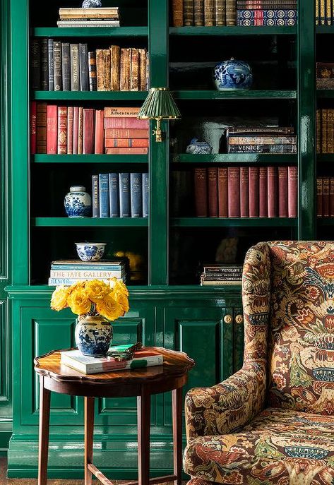 Living room features a glossy emerald green built-in bookcase with cabinets that boast brass knobs, a brown flocked wingback chair and a round wooden accent table. Green Built In Bookcase, Bookshelf Color Ideas Paint, Timeless Homes, Calke Green, Apartment Refresh, Green Bookshelves, Academia Library, Green Library, Bookcase Ideas