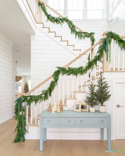 A coastal Christmas home entryway with white oak hardwood floors, white shiplap walls, a blue linen console table, garland on the staircase, and neutral and blue accents. Coastal Christmas Staircase, Coastal Christmas Wedding, Coastal Christmas Porch Decor, Christmas In The Hamptons, Christmas Coastal Decor, Lake Christmas Decor, Coastal Holiday Decor, Lake House Christmas, Nantucket Christmas