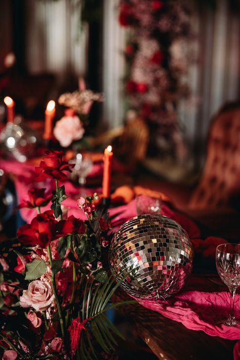 This shows a close up of a colourful top table set up. The colour theme consists of different shades of pinks and reds.On the table is a hot pink muslin table runner, orange napkins, different sized disco balls, floral arrangements and black candlesticks with orange candles. Colorful Disco Ball Wedding, Flowers And Disco Ball Party, Jewel Tone Disco Wedding, Colourful Disco Wedding, Disco Balls At Wedding, Disco Wedding Flowers, Retro Eclectic Wedding, Moody Disco Wedding, Garden Disco Wedding