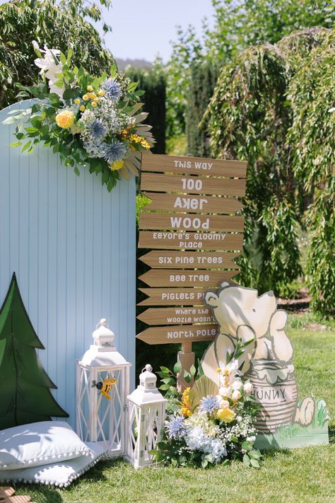 Photo backdrop setup with light blue solid wood arch, blue and yellow flowers, pastel Winnie the Pooh cardboard cutout, white lanterns, and a wooden arrow sign of the 100 Acre Woods. Diy 100 Acre Wood Sign, Blue Winnie The Pooh Baby Shower Ideas, 100 Acre Woods Party, Hundred Acre Woods Baby Shower Ideas, 100 Acre Woods Baby Shower Ideas, Hundred Acre Wood Sign, Winnie The Pooh Wedding, Wooden Arrow Sign, Brit Milah