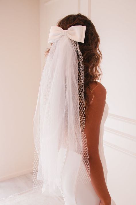 The Ariana Veil is a simplistic bridal veil made with light ivory tulle, English Merry Widow Netting, and topped with a light ivory satin bow. All elements of this veil are attached to a clamp clip to easily secure into your hair. The Ariana Veil is designed to accompany a simplistic bridal look, to wear with a second look at your reception, or to dress up any bridal festivity outfit. Details: Color: Light Ivory Bow Dimensions: 7.5 x 3 inches Tulle Length: 30 inches Netting Length: 27 inches All Wedding Hair With No Veil, Slick Back Bun With Veil, Bridal Hair With Bow Veil, Tie Sleeve Wedding Dress, Wedding Veil Bow, Veil Designs Wedding, Wedding Bow Hair, Bow Veil Wedding, Bow Veils