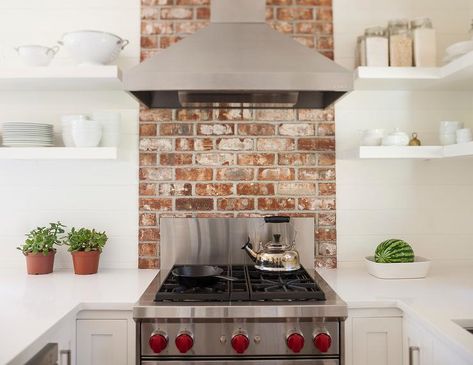 Stainless steel vent hood and wolf range unites with a red brick cooktop backsplash flanked by white floating shelves, white subway tiles, and white quartz countertops. Cottage Backsplash, Red Brick Backsplash, Stove Backsplash Ideas, Cooktop Backsplash, Brick Slips Kitchen, Red Brick Kitchen, Brick Tiles Kitchen, Brick Stove, Kitchen Extras