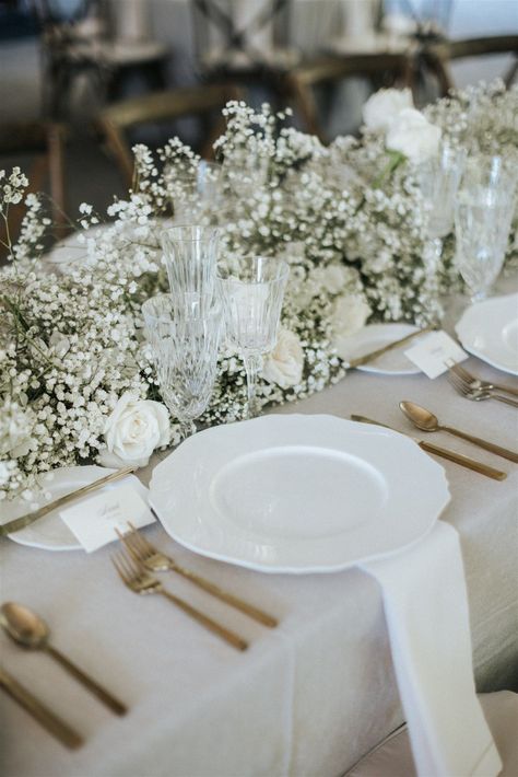 Image shows a closeup of a wedding table for guests. The linen is a neutral beige color with gold sileverware and a white plate setting. The centerpieces is a beautiful and simple floral arrangement that runs along the table of baby's breath and white roses. Babies Breath Wedding, White Rose Centerpieces, Engagement Party Table, Wedding Planning Boards, Bridal Party Tables, Black And White Wedding Theme, Table Arrangements Wedding, Minimalist Wedding Decor, Enchanting Wedding