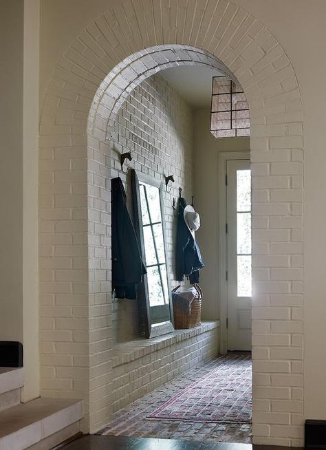 A white brick arched mudroom door way opens to red brick floor tiles covered in a red rug placed in front of a built-in white brick bench topped with a full length mirror leaning against a white brick wall between two iron horse coat hooks. Brick Mudroom, Brick Wall Ideas, Arch Entryway, White Brick Wallpaper, Brick Archway, Brick Floor, Interior Brick, Brick Interior, Look Wallpaper