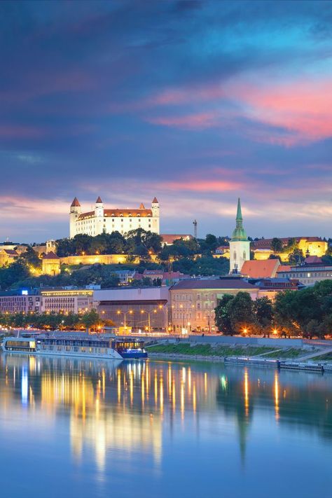 The text “Bratislava Travel Guide: A Reveal of Slovakia’s Beautiful Capital” overlaying a photo of Bratislava Castle lit-up under a purple and blue night sky, its reflection vivid in the beautiful blue Danube River Bratislava Slovakia, European City, Brno, Secret Places, Central Europe, Bratislava, Capital City, Slovakia, Holiday Travel