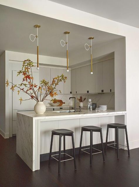 Kitchen features backless black stools at a honed marble waterfall edge peninsula illuminated by white and gold wand lights and light gray cabinets with black pulls. Waterfall Peninsula Kitchen, Waterfall Peninsula, White Beadboard Kitchen, Kitchen Peninsula Design, White Kitchen Peninsula, Peninsula Kitchen Design, Flat Front Cabinets, Black Pulls, Light Grey Kitchen Cabinets