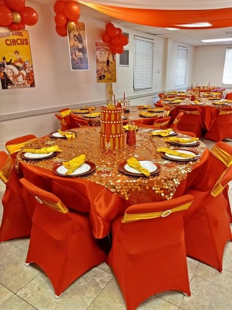 Add imperial charm to your circus themed party décor by dressing your tables with orange tablecloths and gold sequin overlays. Create a presentable place setting by arranging yellow napkins with orange chargers and white plates as well as opt for red taper candles to enhance your display. Also, adorn your chairs with orange covers and gold sashes and decorate your empty walls with gold and orange balloons. Lastly, cover your ceiling with orange and white drapes for a cohesive appeal. Red And Yellow Table Setting, Orange And Gold Decorations, Orange And Yellow Party Decorations, Iftaar Ideas, Red Taper Candles, Yellow Party Decorations, Yellow Dinner, 75 Birthday, Circus Themed Party