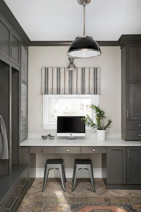 This well styled mudroom and home office combo features silver Tolix stools placed on red brick herringbone pavers beneath a gray oak built-in desk accented with satin nickel hardware and a white quartz countertop. Boot Room And Office, Mud Room And Office Combo, Mudroom With Desk Area, Mudroom And Office Combo, Boot Room Office, Office Mudroom Combo, Desk In Mudroom, Mud Room Office Combo, Mudroom With Desk