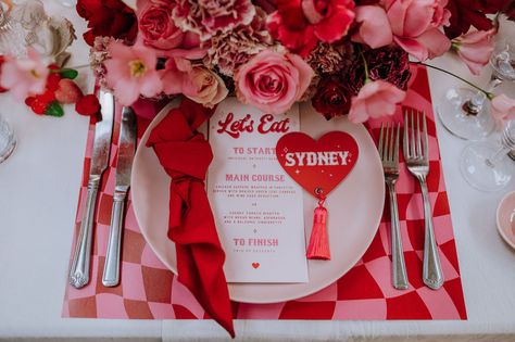 A table setting with red and pink checkerboard placemat, a pink plate with a red napkin, menu and red heart shaped name card with a red tassel with red and pink flowers at the top of the image Pink And Red Retro Wedding, Pink Red Table Setting, Valentine Wedding Theme, Valentines Themed Wedding, Pink And Red Table Setting, Valentines Wedding Theme, Valentine’s Day Wedding, Valentines Theme Wedding, Pink And Red Wedding Theme