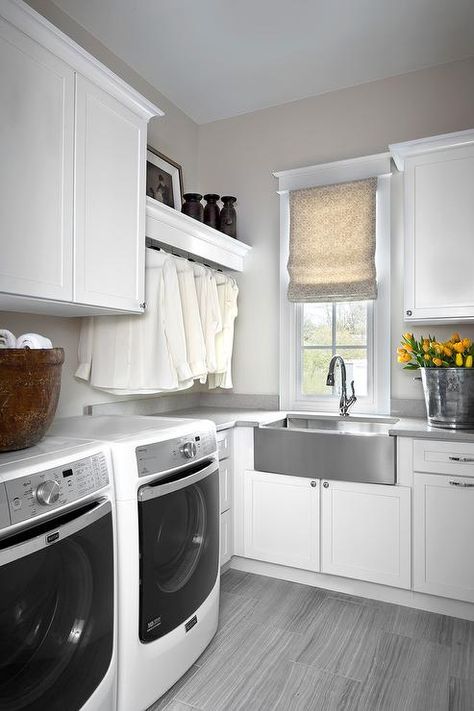 Stunning gray and white transitional laundry room with gray wood like tiled floors is fitted with a stainless steel apron sink complimented with a polished nickel gooseneck faucet positioned under a window dressed in a beige roman shade framed by light gray walls accenting gray quartz countertops sat atop white cabinets. Fold Out Ironing Board, Dog Sink, Gray Wood Floors, Tile Around Bathtub, Remodel Pantry, Stainless Steel Apron Sink, Laundry Room Idea, Gray Quartz Countertops, Transitional Laundry Room