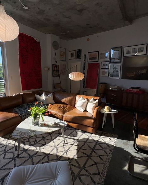 Photo of a modern living room with a brown leather couch, a white coffee table and red walls. Sunlight is coming in from large windows. The ceiling is concrete. There is a gallery wall with framed pictures. Warm lighting gives the room a moody, modern style. A white rug and black furniture complete the look. Brown Leather Couch Aesthetic, Leather Couch Apartment, Leather Sofa Living, Brown Leather Couch, Mid Century Modern Living Room, Leather Couch, Black Furniture, Red Walls, Best Sofa