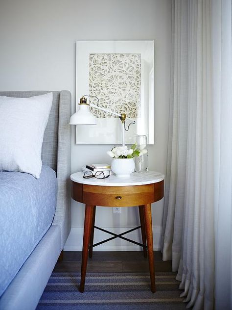 Restful white, gray, and blue bedroom boasts a heather gray wingback bed covered in white and blue bedding and positioned beside a West Elm Penelope Nightstand illuminated by a white swing arm table lamp placed in front of a white framed art piece beside a windows dressed in white curtains. Dining Photoshoot, Chair Nightstand, Gray Upholstered Bed, Round Nightstand, Restful Bedrooms, Marble Tables Design, Marble House, Wingback Bed, Bedroom White