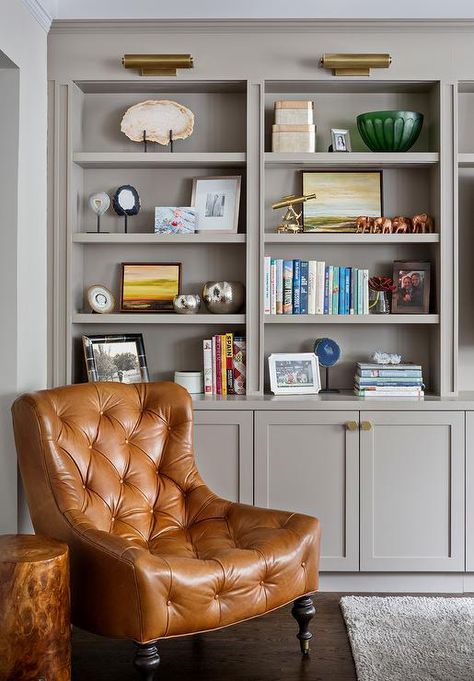 Cognac leather tufted corner reading chair is positioned on caster legs in front of built in styled gray bookshelves located above gray shaker TV cabinets fitted with brass hexagon knobs lit by brass picture lights. Grey Bookshelves, Styling Bookshelves, Leather Chairs, Furniture Placement, Living Room Shelves, Trendy Living Rooms, Transitional Living Rooms, Built In Bookcase, New Living Room